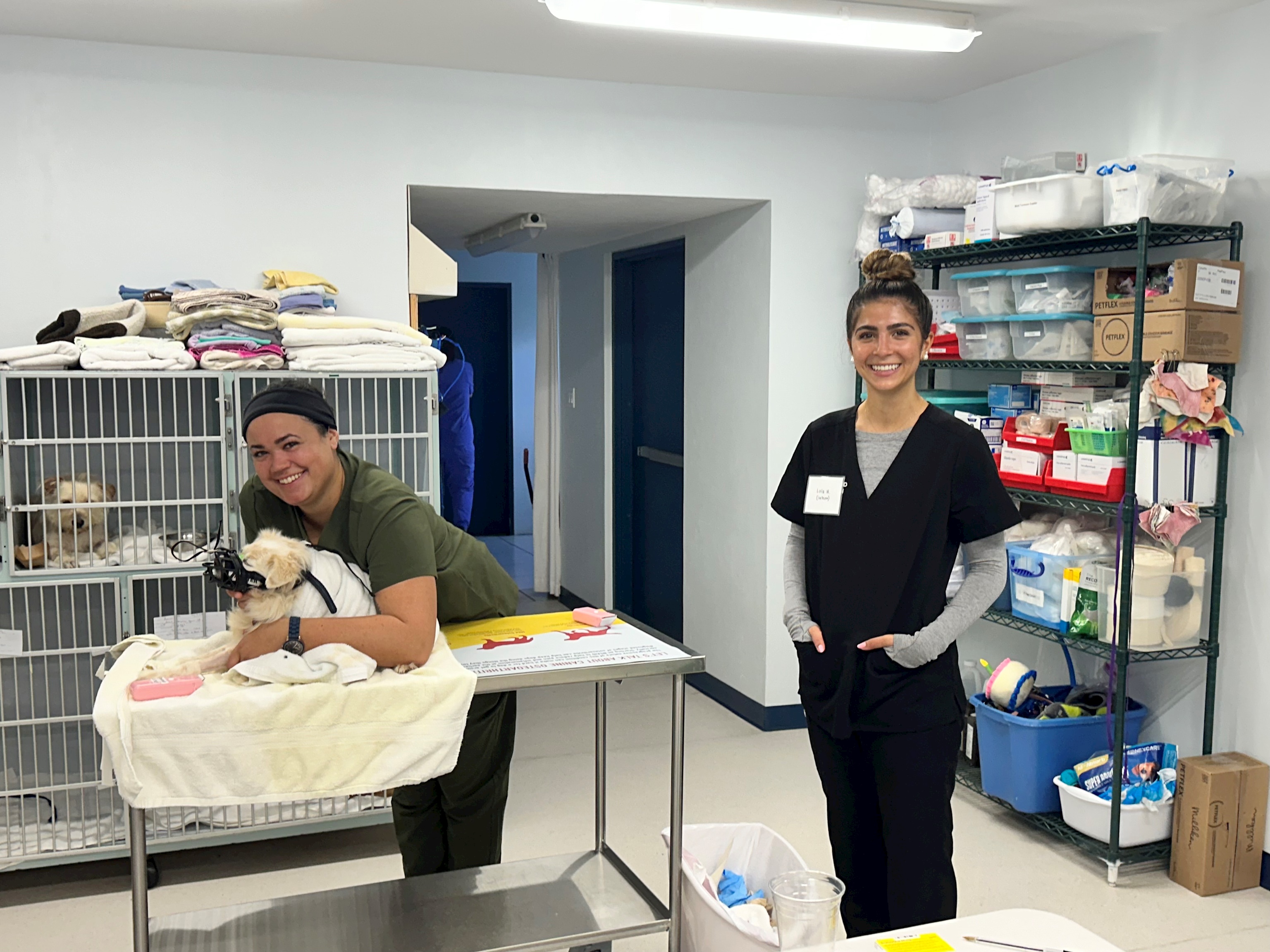 Lola wears scrubs and smiles in the back portion of a vet clinic. A vet tech handles a dog on a metal table next to Lola.